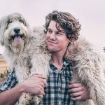 A man carrying his white scruffy dog on his shoulders at a farm.