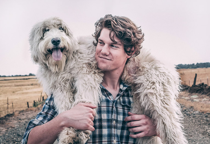 A man carrying his white scruffy dog on his shoulders at a farm.
